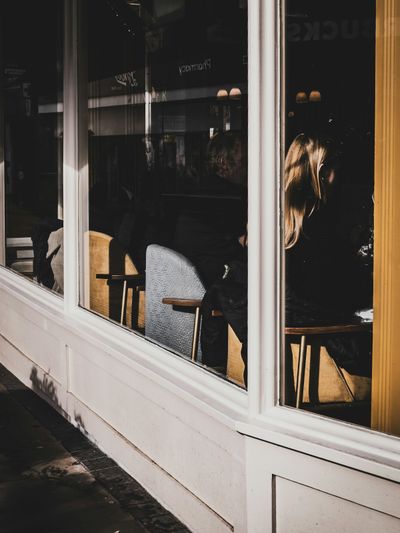 The cafe exterior corner seen through a large glass window, a woman's silhouette visible inside against warm light