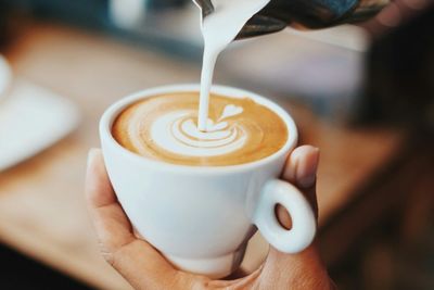 A hand pouring cream over a coffee in a ceramic cup